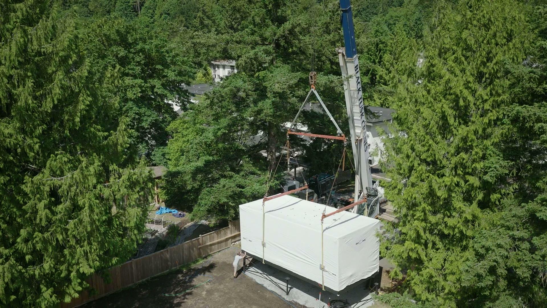 Wrapped ADU module hanging from a crane over a pad in the trees